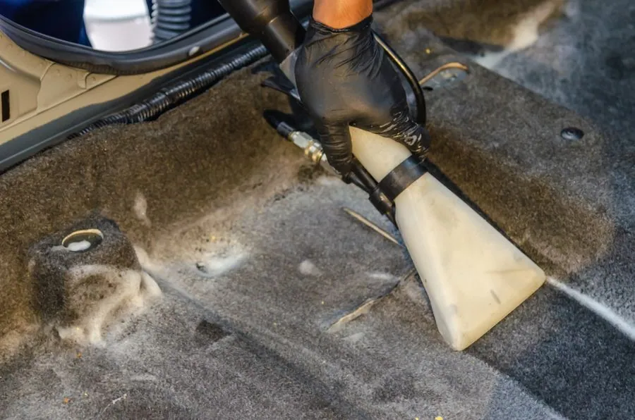 Hand im schwarzen Handschuh reinigt den Teppichboden im Fahrzeug mit der Düse eines Waschsaugers